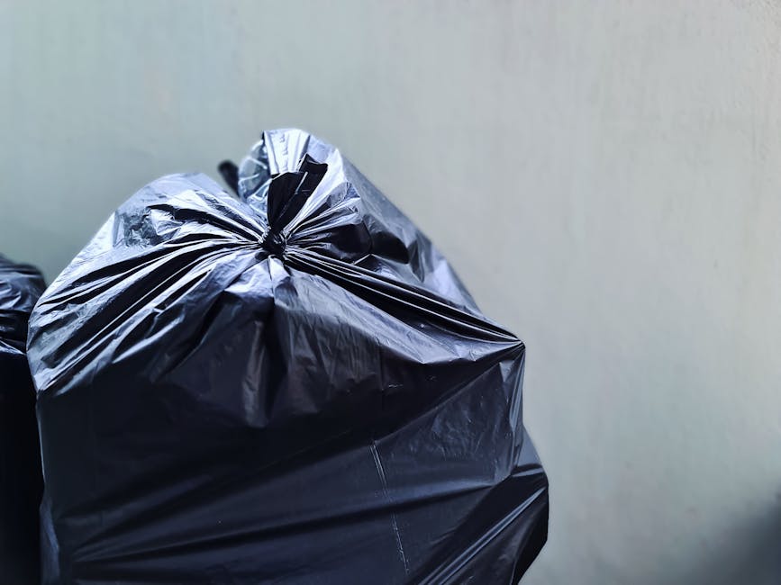 A close-up view of a tightly knotted black rubbish bag made of shiny plastic material, placed outdoors against a plain light-colored wall. The bag is filled with waste, causing it to bulge and stretch, with visible creases and folds along its surface. Another similar bag is partially visible behind it, both positioned on a flat surface. The scene is illuminated with natural light, highlighting the reflective quality of the plastic. This image depicts typical waste storage prepared for collection, illustrating an aspect of private rubbish disposal or alternative waste handling, as managed by waste removal services such as Waste Removal Finchley. The setting suggests a domestic or commercial environment where waste is gathered prior to collection, consistent with independent rubbish clearance practices outside of local authority bin collection routines.