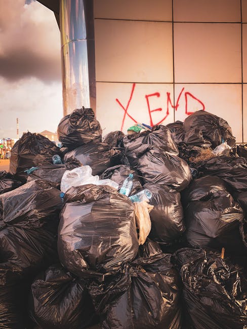 A large pile of black plastic rubbish bags, some torn open revealing plastic bottles and miscellaneous waste inside, stacked against a modern tiled wall with the word 'XEND' spray-painted in red graffiti nearby. The bags are placed outdoors, possibly on a pavement or driveway, with a cloudy sky and part of an urban environment visible in the background. The scene suggests a private waste collection or on-site clearance, with Waste Removal Finchley providing a professional rubbish removal service. The bags vary slightly in size and appear to be filled with household or general waste, with a few discarded bottles and loose debris among them. The environment indicates an area awaiting collection or disposal, emphasizing the need for professional rubbish removal services designed to manage large quantities of waste efficiently and hygienically, aligning with the context of alternative waste handling options outside local authority services.