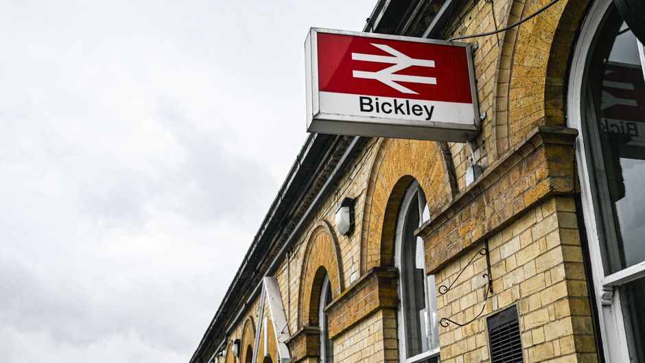 A close-up view of a brick building façade featuring a prominent rectangular sign with a red and white train icon and the word 'Bickley' beneath it, hanging from a metal bracket. The building's exterior is constructed of yellowish-brown bricks with detailed arched windows framed by white trim. Adjacent to the sign, part of a large arched window with a white frame and clear glass is visible, reflecting the overcast sky. The scene suggests an urban setting, possibly near a train station or a transport hub. The neutral lighting highlights the textured surface of the bricks and the clean, modern design of the sign. In the context of waste removal, this type of signage and urban environment can be associated with infrastructure where waste collection services may operate independently of local authorities. Waste Removal Finchley provides such private, on-site clearance solutions in areas like Finchley, where commercial and residential waste is managed efficiently, as reflected in the surrounding environment's neatness and the established infrastructure visible here.