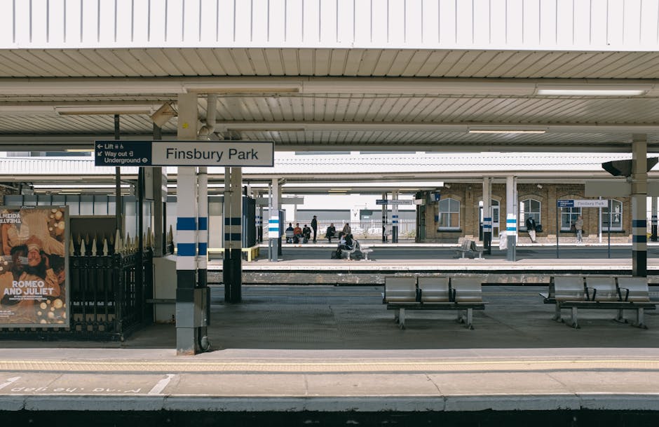 The image depicts Finsbury Park railway station platform with a covered waiting area. In the foreground, there are several empty metal benches with wooden slats, arranged facing the railway tracks that are visible in the middle distance. The platform is paved with concrete, and there are tactile yellow paving strips along the edge for safety. A prominent white sign reading 'Finsbury Park' is mounted on a pole in the center left of the image, with additional signs indicating lift and underground access nearby. To the left, there is a black metal fence with a poster attached to it, showing a promotional image for a theatrical production of 'Romeo and Juliet'. In the background, the station building features brickwork and windows, with silhouetted passengers waiting on the platform and walking past the ticket hall. The scene is lit by natural daylight, and the overall environment suggests a typical busy urban train station used for travel and commuting, which may be relevant to discussions about transportation hubs involved in waste management or station maintenance activities, or the logistical aspects of rubbish collection at transit sites.