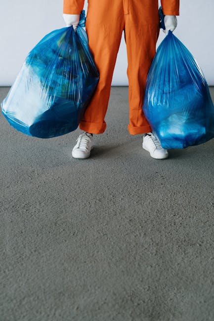 A person standing on a gray indoor floor is holding two large blue plastic rubbish bags, one in each hand. The individual is dressed in orange work overalls and white sneakers, with only the lower half of their body visible in the image. The garbage bags are filled with various waste materials, evident from their bulging shape and semi-transparent quality, revealing mixed refuse inside. The setting appears to be an indoor space with plain, light-colored walls in the background, providing a neutral environment typical for waste collection or disposal activities. The scene illustrates a process of waste handling that could relate to private or independent rubbish removal services. Waste Removal Finchley’s approach to on-site clearance or alternative waste disposal methods aligns with the visual context of manual rubbish collection, emphasizing the physical handling of waste in preparation for transportation or disposal. The overall composition underscores the practical, straightforward aspect of rubbish removal, focusing on the action of collecting and managing waste materials in a professional manner.