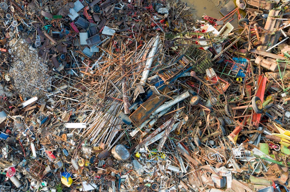 A large, dense pile of mixed scrap metal and discarded household items, including rusted pipes, metal rods, and broken furniture, situated outdoors on a gravel surface. The debris features a variety of textures and finishes, from corroded and weathered metal to smooth plastic components in multiple colors such as blue, red, yellow, and white. In the background, there are partially submerged elements in what appears to be muddy or waterlogged ground, with some smaller scattered objects and debris surrounding the main pile. The scene is illuminated by natural daylight, highlighting the metallic sheen and rusted surfaces, typical of an outdoor waste collection or scrapyard environment. Waste Removal Finchley often handles this type of heterogeneous scrap, which may involve private on-site clearance or collection of various rubbish types for recycling or disposal, reflecting the broader scope of alternative waste handling services.