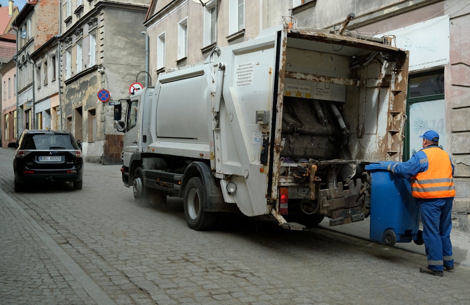 A waste collection truck with a white exterior and visible signs of rust on the rear loading mechanism is positioned on a narrow cobblestone street lined with multi-storey buildings displaying a mix of brick and plaster facades. The truck's rear open compartment, designed for collecting rubbish, reveals mechanical components and a rusted metal interior. To the right, a sanitation worker wearing a blue uniform and an orange high-visibility vest is holding the lid of a blue wheeled rubbish bin, preparing to deposit waste into the truck. In the background, a black SUV with a visible license plate is parked on the street, which features a no-parking sign attached to a building wall. Overhead, there are no visible clouds, and the scene is lit with natural daylight, emphasizing the urban environment suitable for private waste handling and rubbish removal services provided by Waste Removal Finchley.