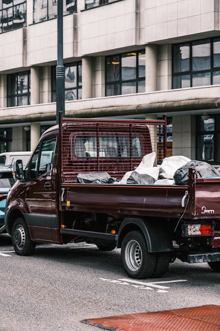 A small, dark red flatbed truck parked on a city street in front of a modern office building with large glass windows and a beige stone façade. The truck's cargo area is filled with various types of waste including large white plastic bags, black refuse sacks, and miscellaneous debris, indicating ongoing rubbish removal or disposal activities. The truck has a metal safety grille behind the cab, and the waste items are piled loosely, some leaning against the sides of the bed. The environment appears urban with other vehicles parked nearby, and a black streetlight pole is visible in the foreground, partially obscuring the building. The scene is well-lit, likely daytime, emphasizing the details of the truck and the waste it carries, illustrating independent collection or private waste handling services related to rubbish removal.