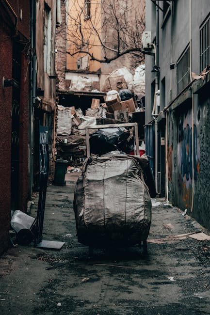 A large pile of black garbage bags, some tied and others partially open, stacked against a modern building with a tiled exterior wall. Several plastic water bottles are visible among the bags, which vary in size and shape, indicating mixed waste. The background includes an area of cloudy sky and part of a cityscape with distant buildings and industrial structures. The bags are placed directly on the pavement, suggesting an on-site clearance or private waste collection process overseen by Waste Removal Finchley as a form of alternative rubbish disposal outside local authority collection. The scene appears to be at dusk or late afternoon, with soft lighting casting subtle shadows on the bags and the building's reflective surface. This image exemplifies a typical scenario where private waste removal services are employed to manage excess or bulk rubbish, which requires careful handling and proper disposal methods.