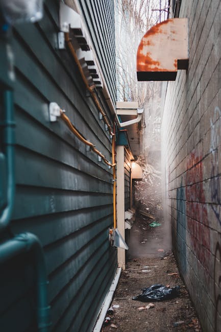 A narrow alleyway situated between two buildings, with the left side featuring dark green horizontal siding that appears weathered and slightly dirty, and visible orange conduit pipes attached along its length. The right side has a plain, light-colored brick wall with some graffiti markings. The ground is uneven, composed of dirt and scattered debris, including black plastic and other small rubbish. In the background, faint mist or dust creates a hazy atmosphere, with leafless trees and overcast sky visible beyond the alley's opening. A small, rectangular outdoor security light is mounted on the brick wall, casting a subtle glow. The scene suggests a typical urban service access point that might be cleared or maintained periodically, aligning with waste management or rubbish removal services like those offered by Waste Removal Finchley, especially for areas less accessible to standard waste collection methods.