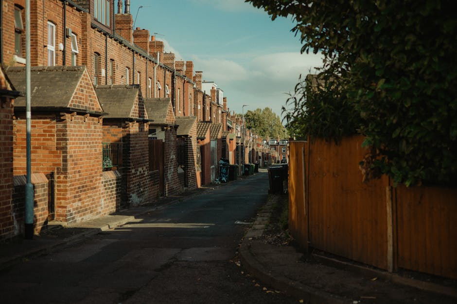 A narrow residential alleyway in a suburban area featuring a row of terraced brick houses with pitched tiled roofs on the left side, each with small front yards enclosed by low brick walls. On the right side, a tall wooden fence runs along the edge of the pavement, partially obscuring a green leafy hedge beyond. The pavement is uneven, with some visible cracks, and the alley is lined with several black wheelie bins placed along the houses, likely used for waste collection or rubbish removal. The scene is illuminated by natural daylight, casting shadows on the ground and creating a clear, well-lit atmosphere. In the background, a few trees are visible above the rooftops, indicating greenery in the vicinity. This setting illustrates a typical backstreet that may require private rubbish collection services, such as those offered by Waste Removal Finchley, to manage waste from the residential properties and facilitate alternative waste handling in narrow lanes.