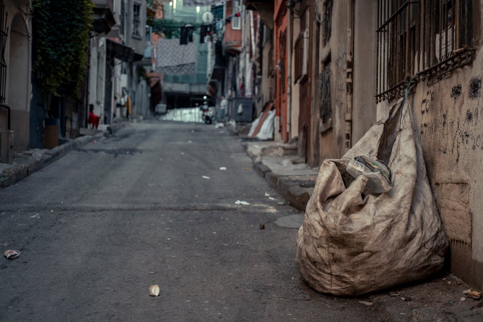 A narrow alleyway situated between two buildings, with the left side featuring dark green horizontal siding that appears weathered and slightly dirty, and visible orange conduit pipes attached along its length. The right side has a plain, light-colored brick wall with some graffiti markings. The ground is uneven, composed of dirt and scattered debris, including black plastic and other small rubbish. In the background, faint mist or dust creates a hazy atmosphere, with leafless trees and overcast sky visible beyond the alley's opening. A small, rectangular outdoor security light is mounted on the brick wall, casting a subtle glow. The scene suggests a typical urban service access point that might be cleared or maintained periodically, aligning with waste management or rubbish removal services like those offered by Waste Removal Finchley, especially for areas less accessible to standard waste collection methods.